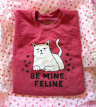 Folded pink shirt with a cat graphic and 'Be Mine, Feline' text on a heart-patterned background